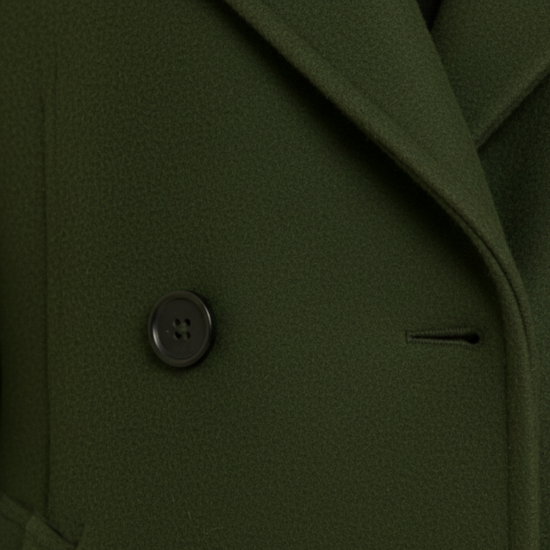 A woman in a knee-length green coat  stands on a cobblestone street, good for cold-weather fashion