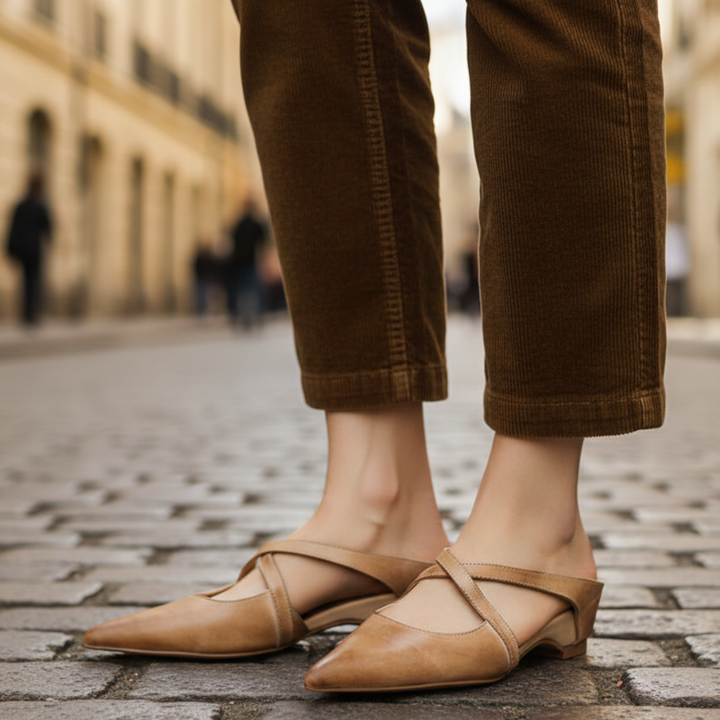A woman's feet in beige leather pointed mule flats with cross-strap detailing, showcasing a modern and elegant style.