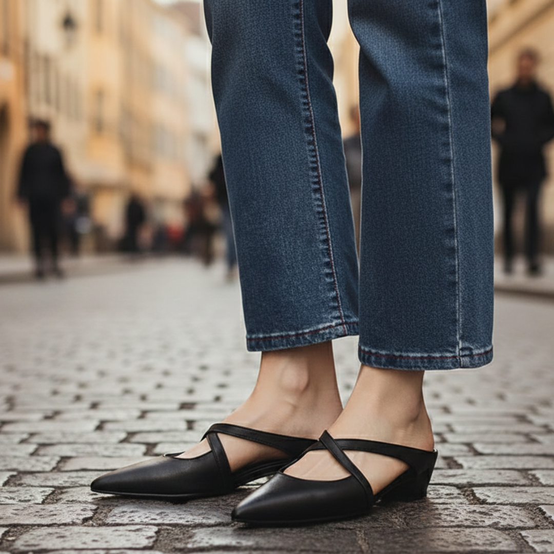 A woman's feet in black leather pointed mule flats with cross-strap detailing, showcasing a modern and elegant style.
